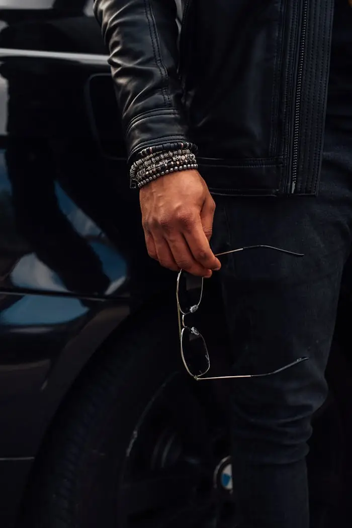 who-we-are Man in leather jacket with bracelets holding sunglasses beside a car.